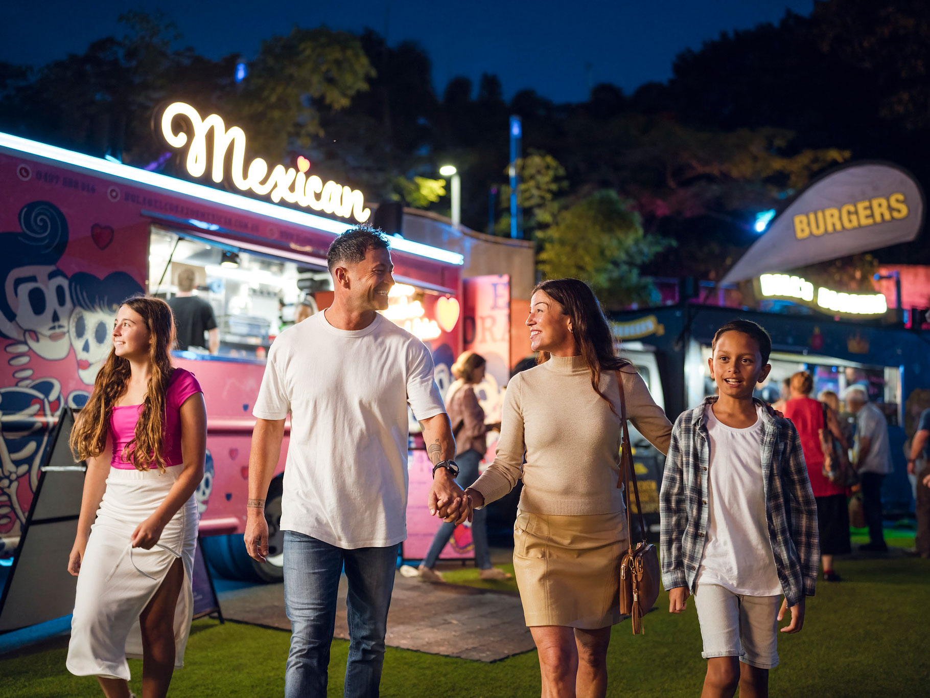 Family of four people walking near food trucks with lights at Celebration Lawn at Roma Street Parkland at night