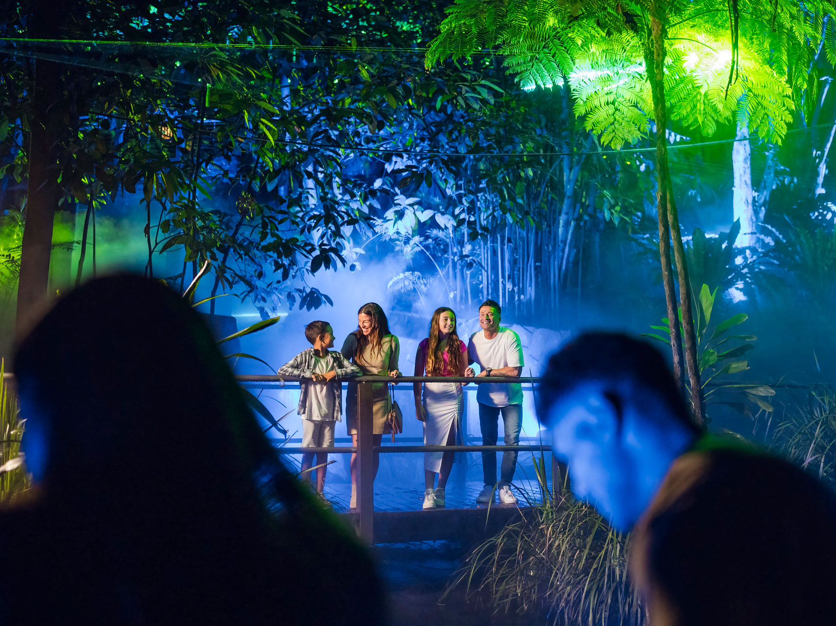 Family of four people standing on an elevated pathway in the rainforest at roma street parkland with colourful ligts on the trees and plants