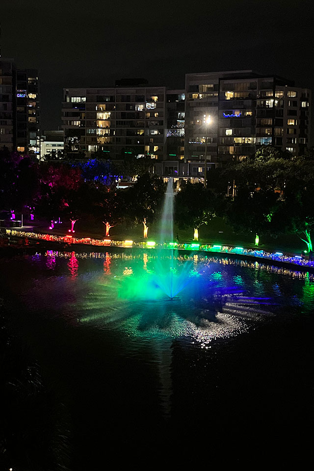 Fountain in the lake lit with rainbow coloured lights and rainbow coloured lights along pathway behind