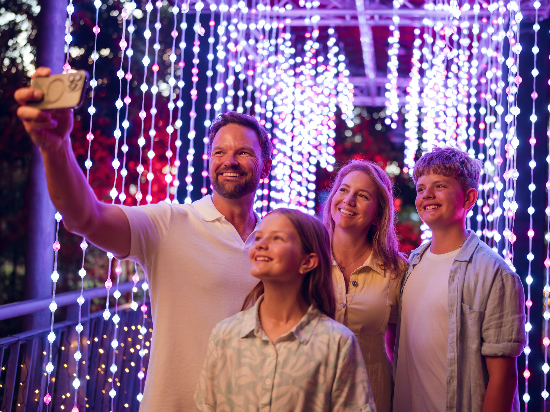 Family of two adults and two children taking a selfie photo on a phone with strings of lights hanging behind them