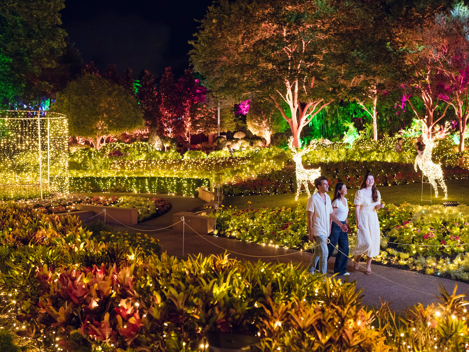 Three friends walking on a path through The Spectacle Garden lit with festive lighting for The Enchanted Garden