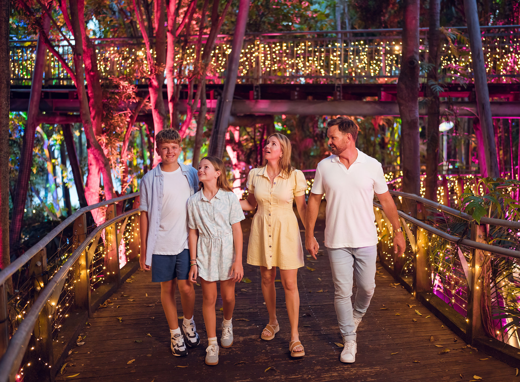 Family including parents and two children walking along a pathway lit with fairy lights for The Enchanted Garden